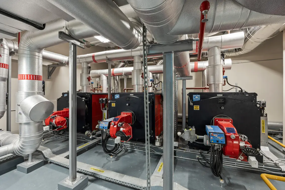 Industrial boiler room with two large black boilers and red burner units amid a network of silver insulated pipes.