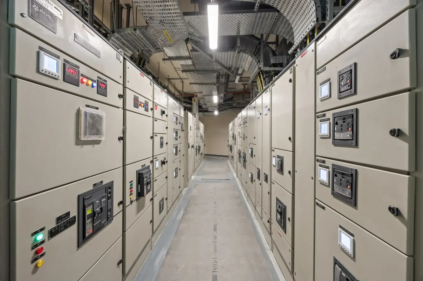 Row of beige electrical switchgear cabinets with control panels along a narrow aisle in a utility room or data center.