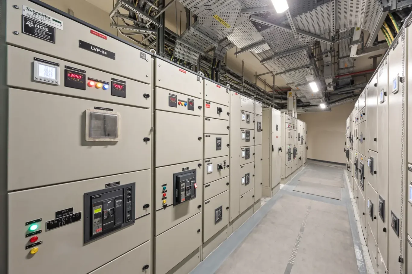 Industrial electrical control room with rows of beige electrical panels and wiring above.