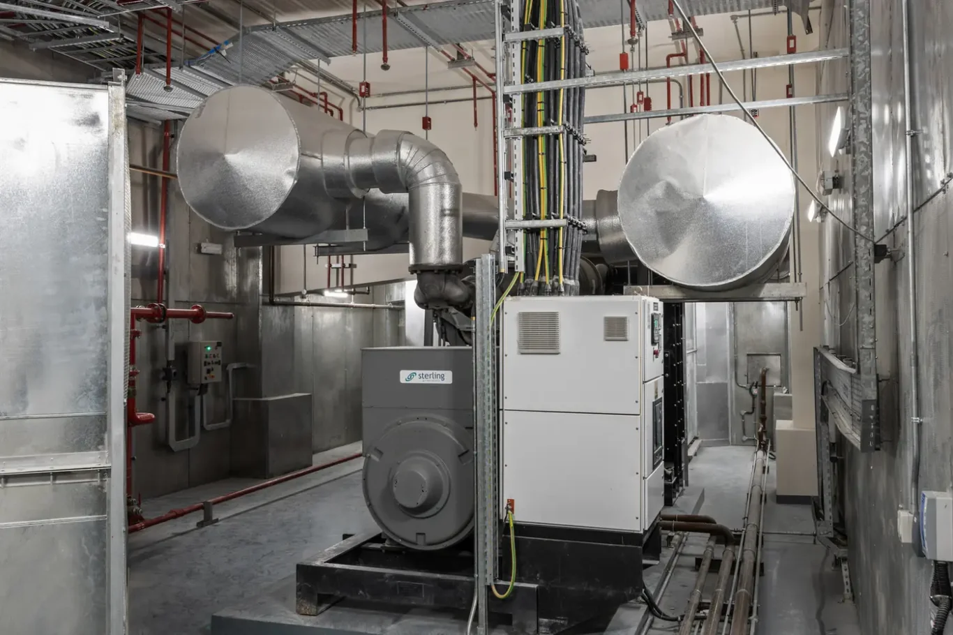 Industrial mechanical room with large silver HVAC ducts, a Sterling-branded unit, and overhead wiring and pipes.