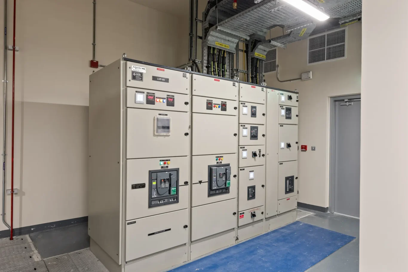 Row of industrial electrical switchgear cabinets with displays and labeled cables in a utilities room.