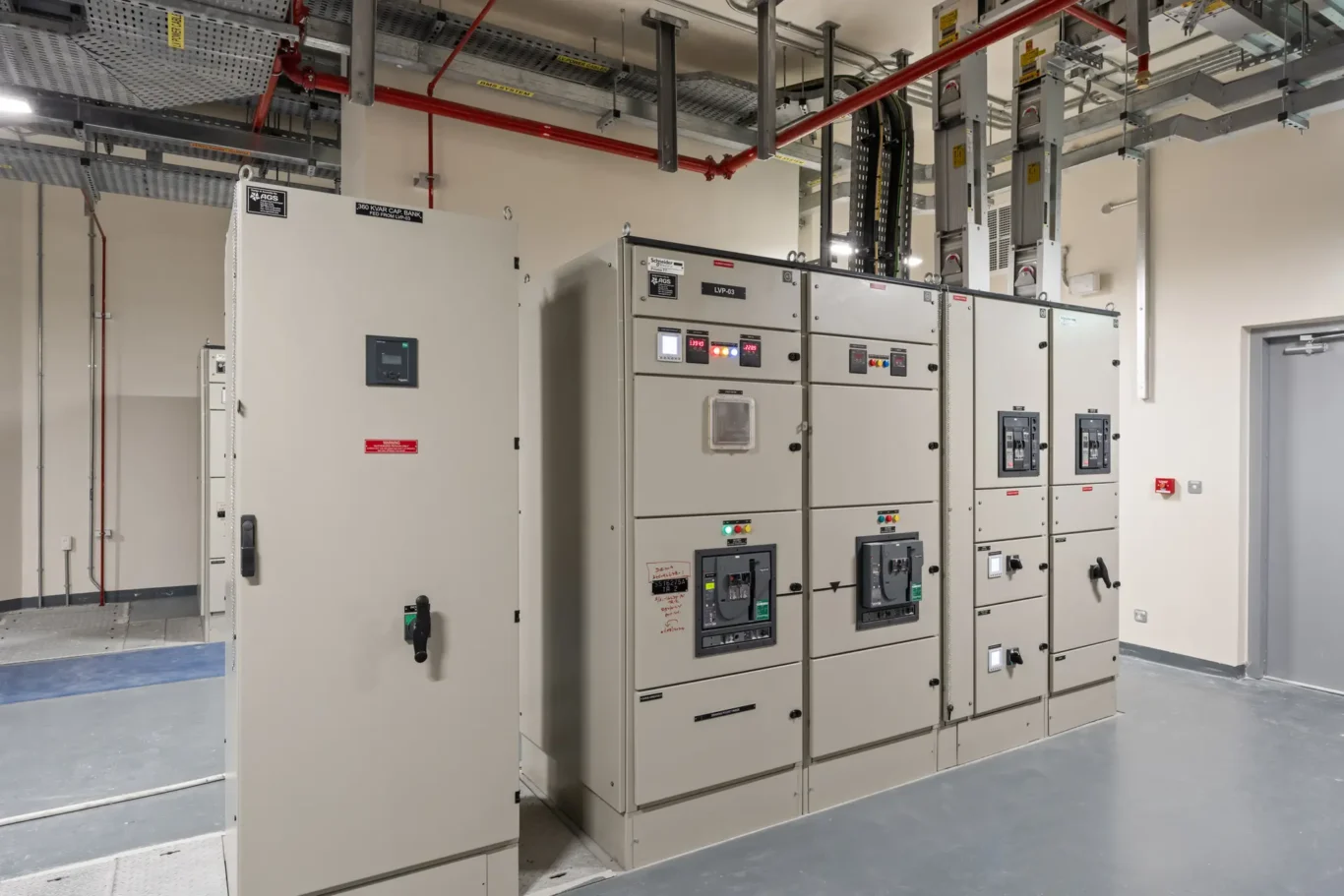 Industrial electrical room with gray switchgear cabinets and control panels under overhead conduit and red pipes.