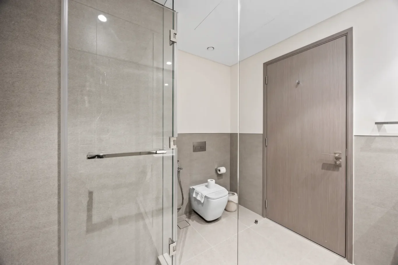 Modern bathroom with a glass shower enclosure on the left, a wall-hung toilet with a folded towel on the lid, and a wooden door on the right.