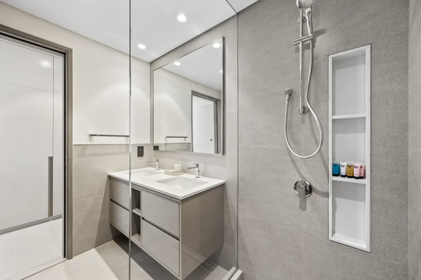 Modern gray bathroom with a double-sink vanity, large mirror, and a glass shower door; built-in vertical niche with assorted toiletries on the right.