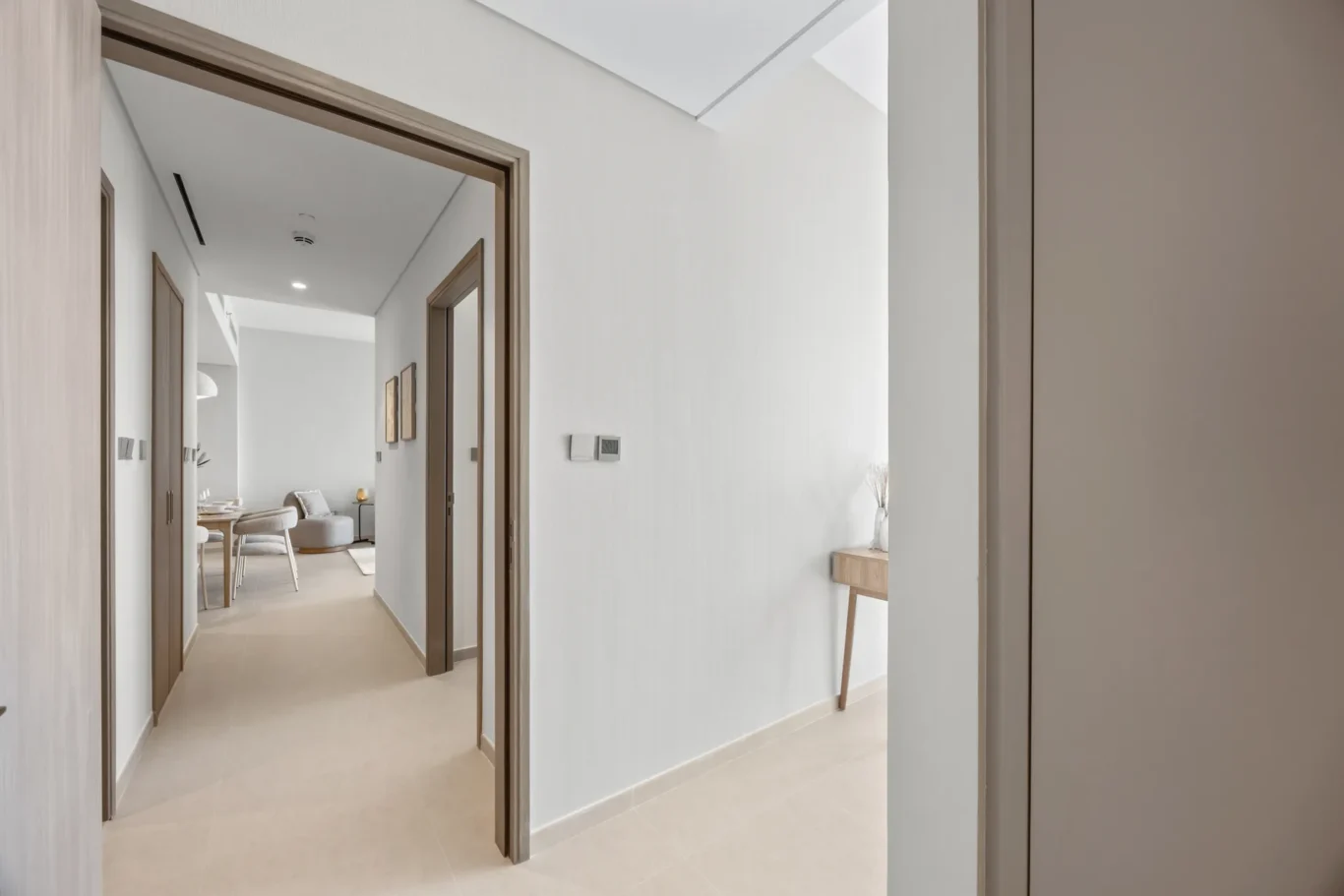 Bright, minimalist hallway with beige tiled floor, white walls, and open doorways leading to a light living/dining area with wooden door frames.