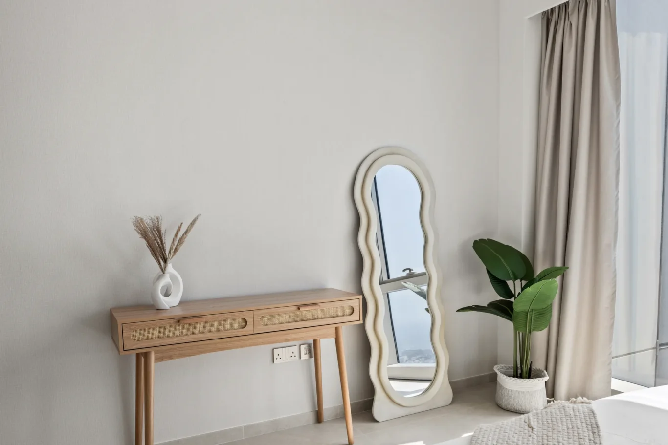 Wavy-framed full-length mirror leaning beside a light wood console table with a vase of dried grasses and a potted plant near beige curtains.