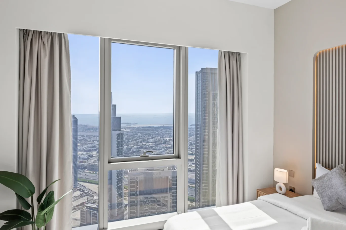 Hotel room with large windows offering a city and sea view, pale curtains, bedside table, lamp, and part of a bed in foreground