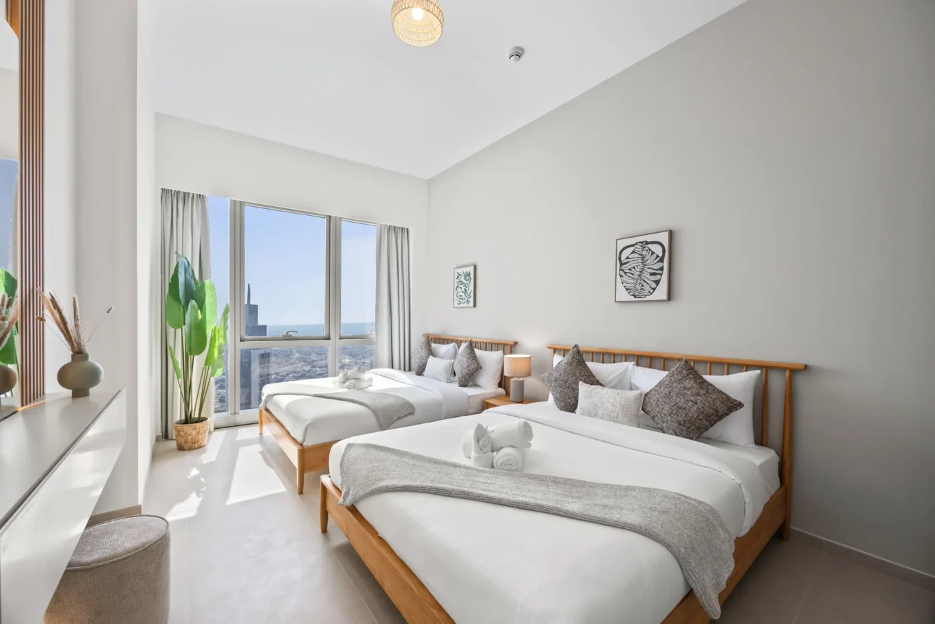 Bright hotel bedroom with two twin beds, white linens, gray throws, wooden headboards, and a large window with a city/ocean view and plants nearby.