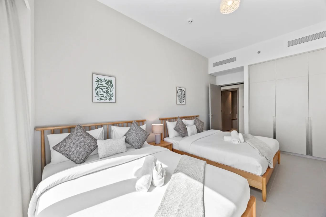 Bright hotel bedroom with two twin beds, white linens, gray pillows, and a wooden headboard; open door and large built-in wardrobe nearby.