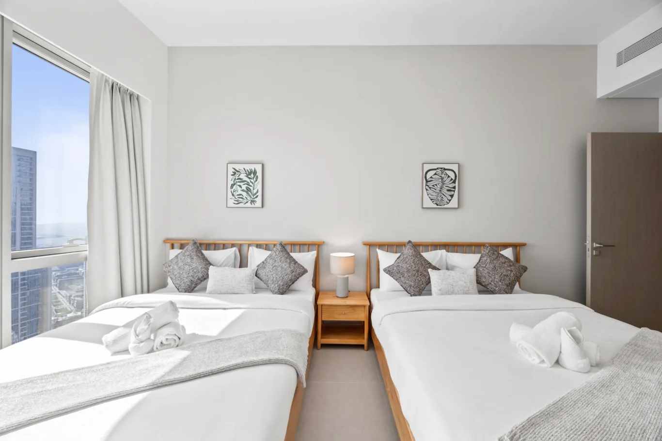 Twin beds with wooden headboards, gray walls, and matching pillows in a bright hotel room with a window view and nightstand between beds.