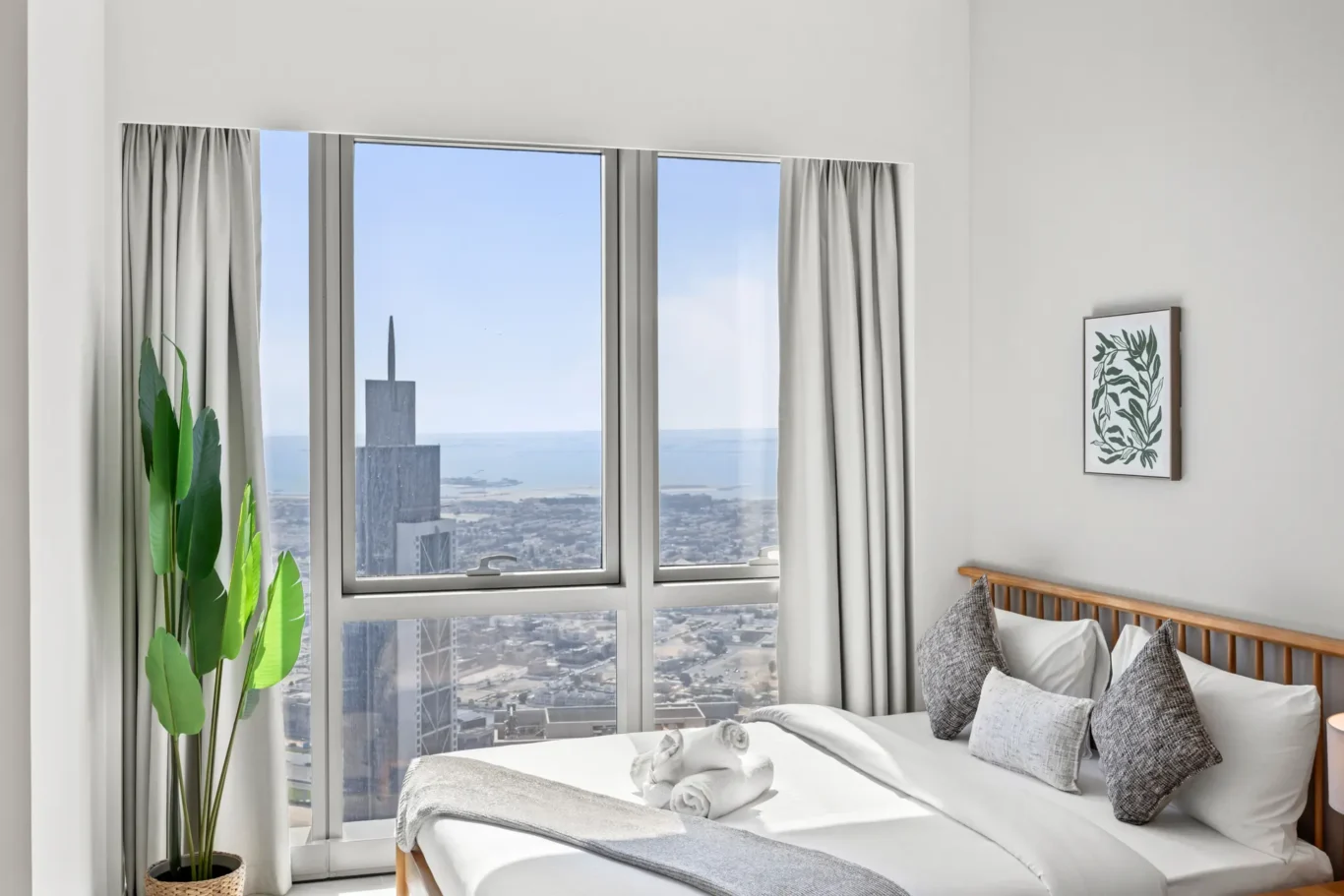 Bedroom with a large window showing a city and ocean view; gray curtains frame the sides and a potted plant sits by the bed.