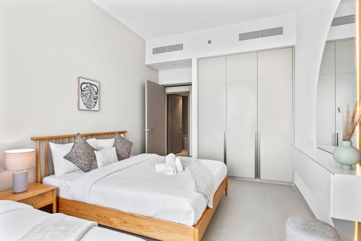 Modern hotel bedroom with a wooden-framed double bed, white bedding, and gray cushions; open door to hallway.