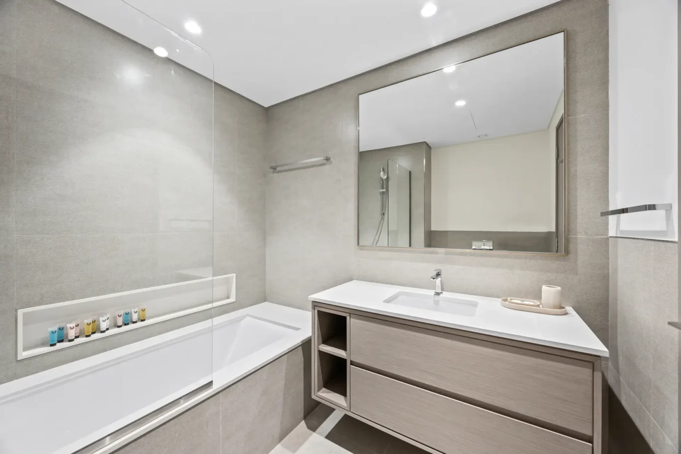 Modern gray-tiled bathroom with a white tub, wall mirror, and wood-finish vanity beneath a large mirror.