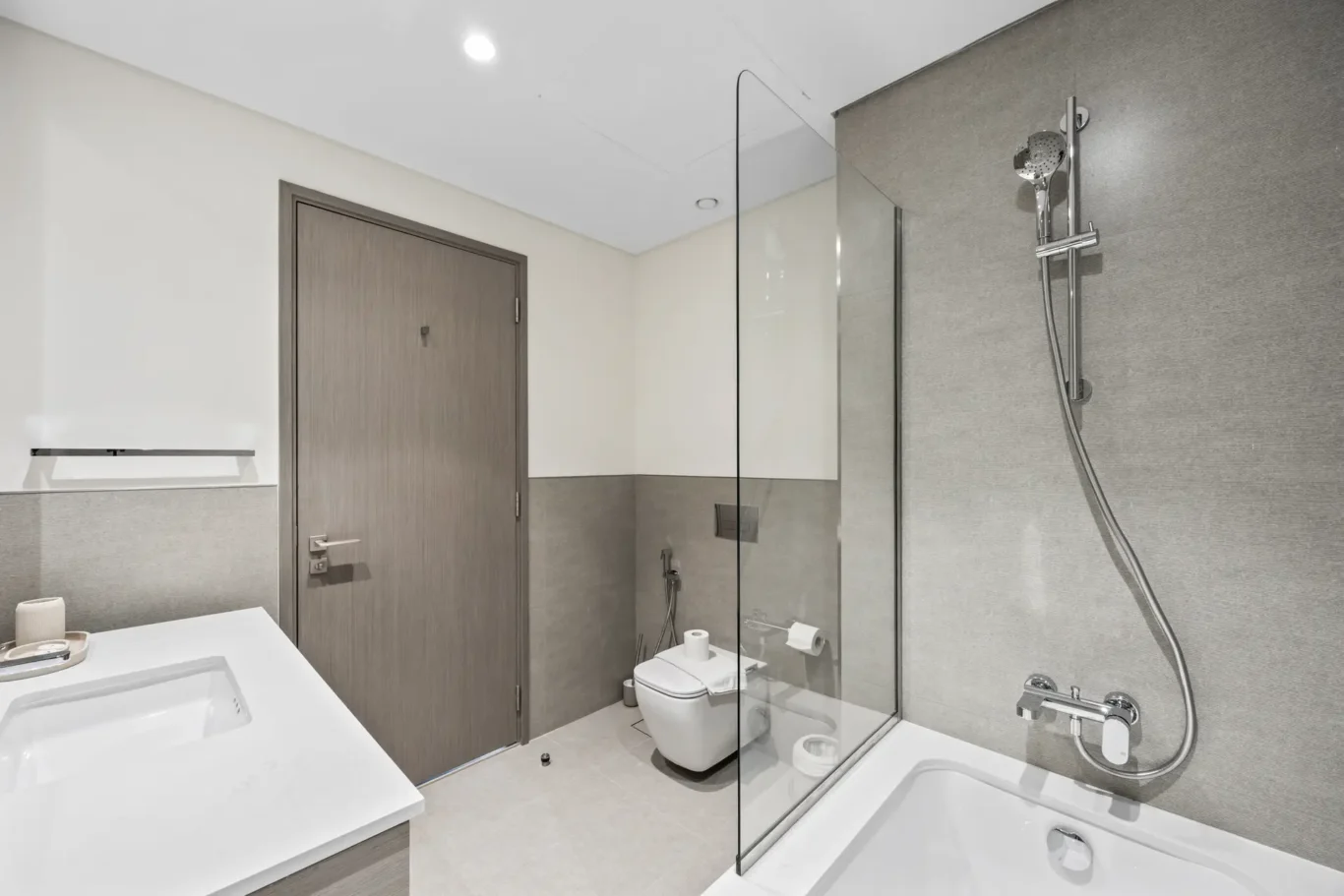 Modern bathroom with glass shower enclosure, wall-hung toilet, and white countertop sink against light walls and a wooden door.