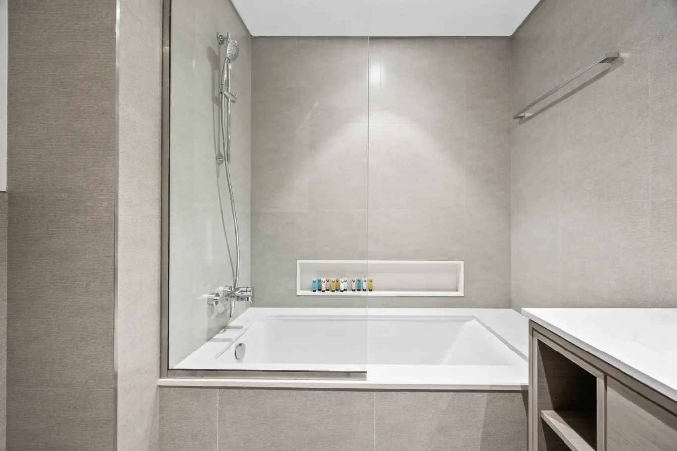 Modern bathroom with glass shower enclosure over a white tub, chrome fixtures, beige tiled walls, and a recessed shelf with small bottles.