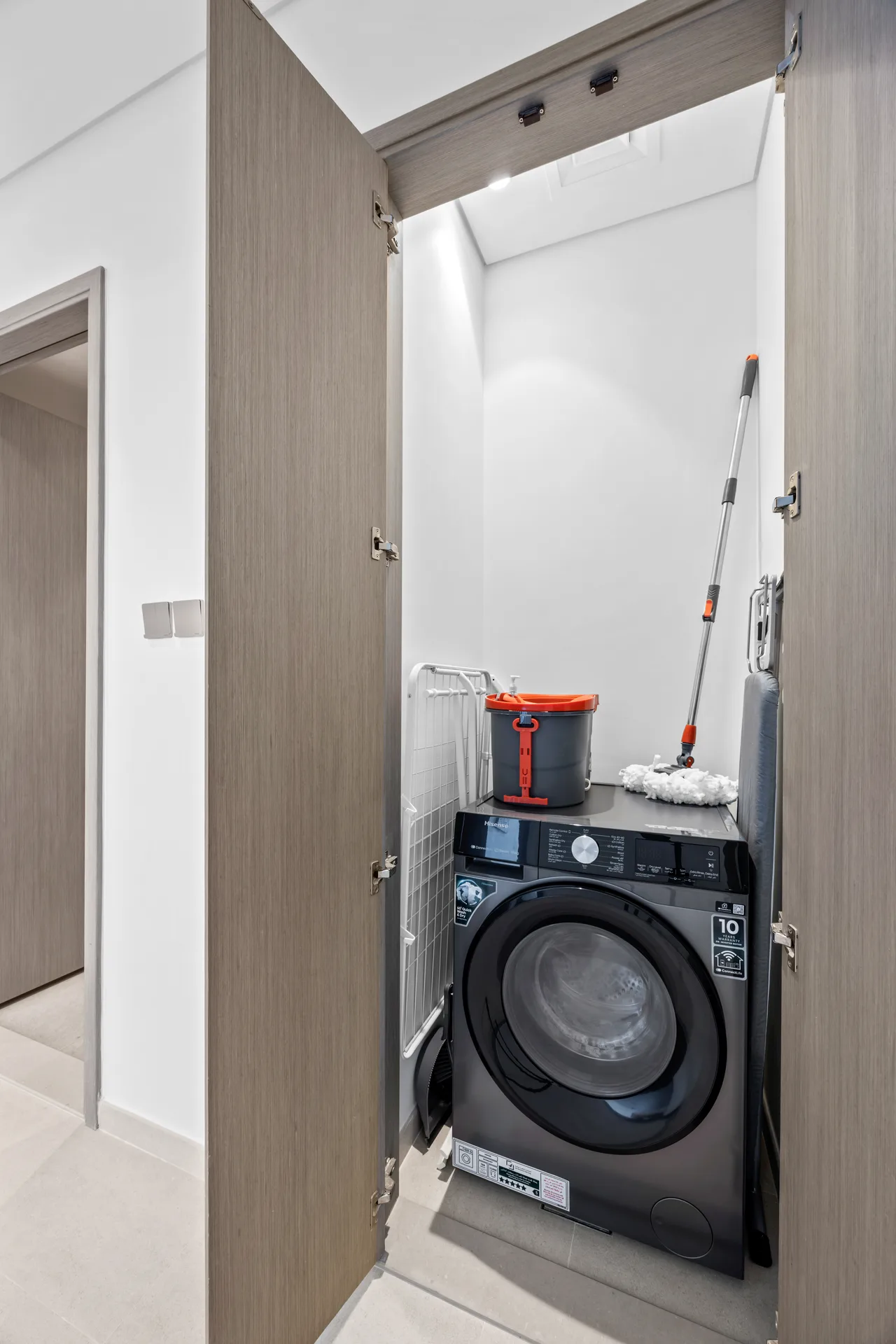 Small laundry closet with a front-loading washing machine, a mop leaning against the wall, and a gray bucket on top of the machine; doors open showing the compact space.
