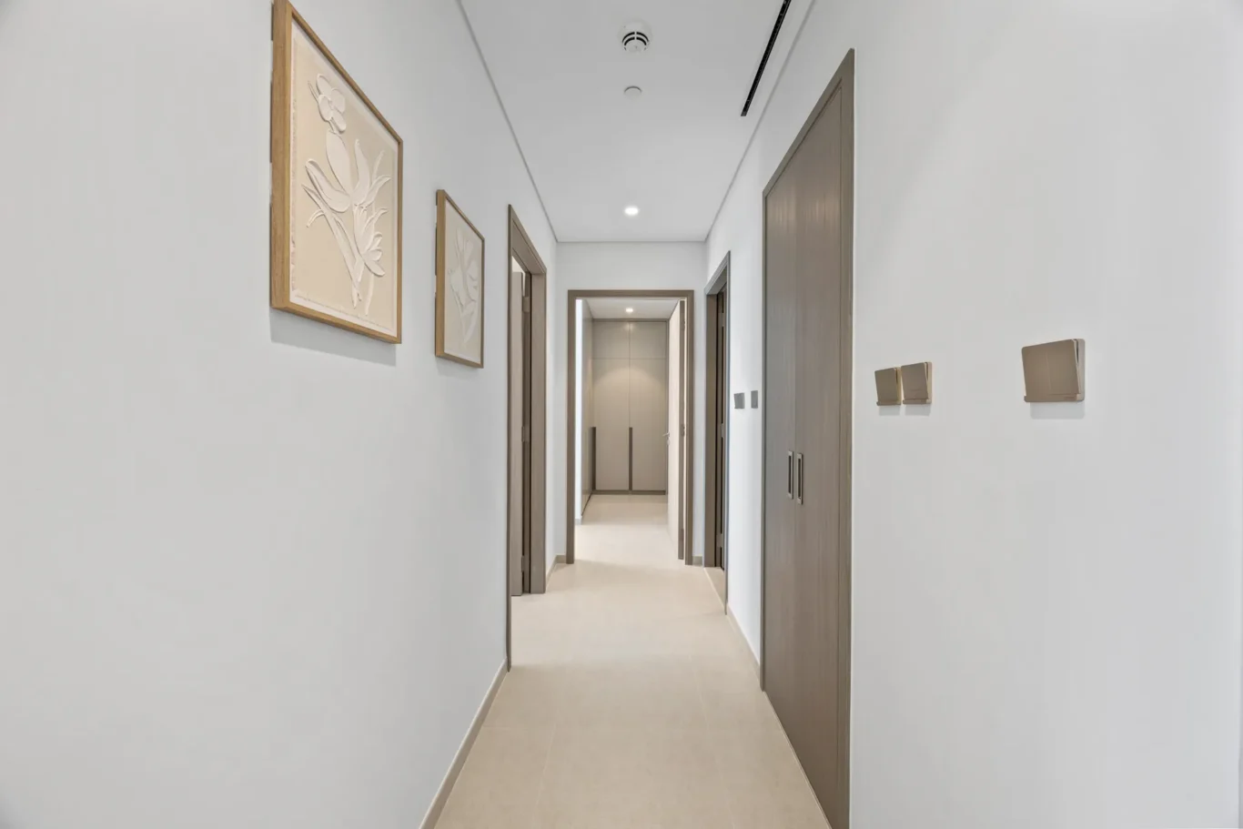 Bright hallway with framed botanical art on the left, beige floor, and an elevator at the far end.
