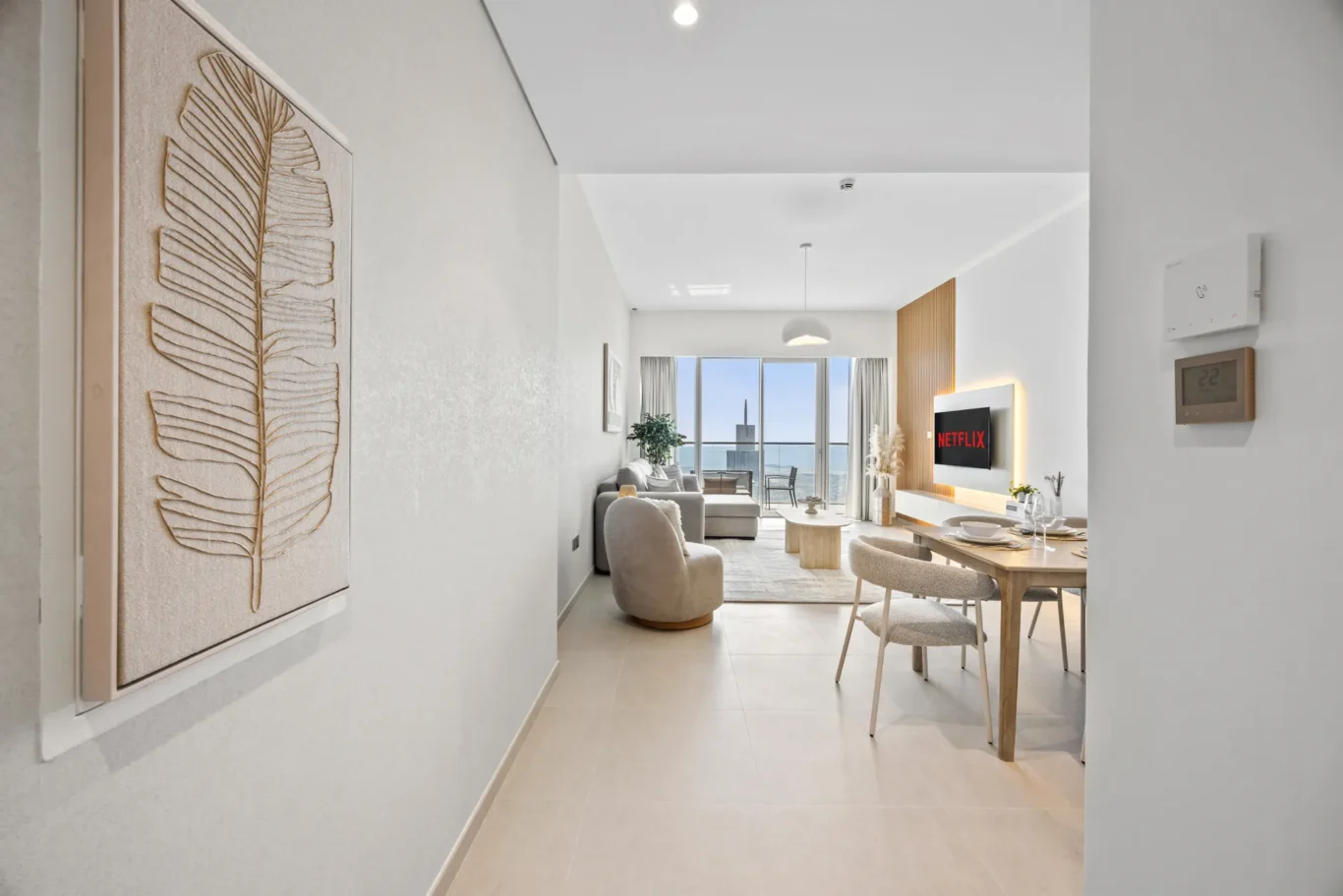 Modern living and dining area with a wall-mounted Netflix TV, a light wood dining table, and a large window balcony with ocean view.