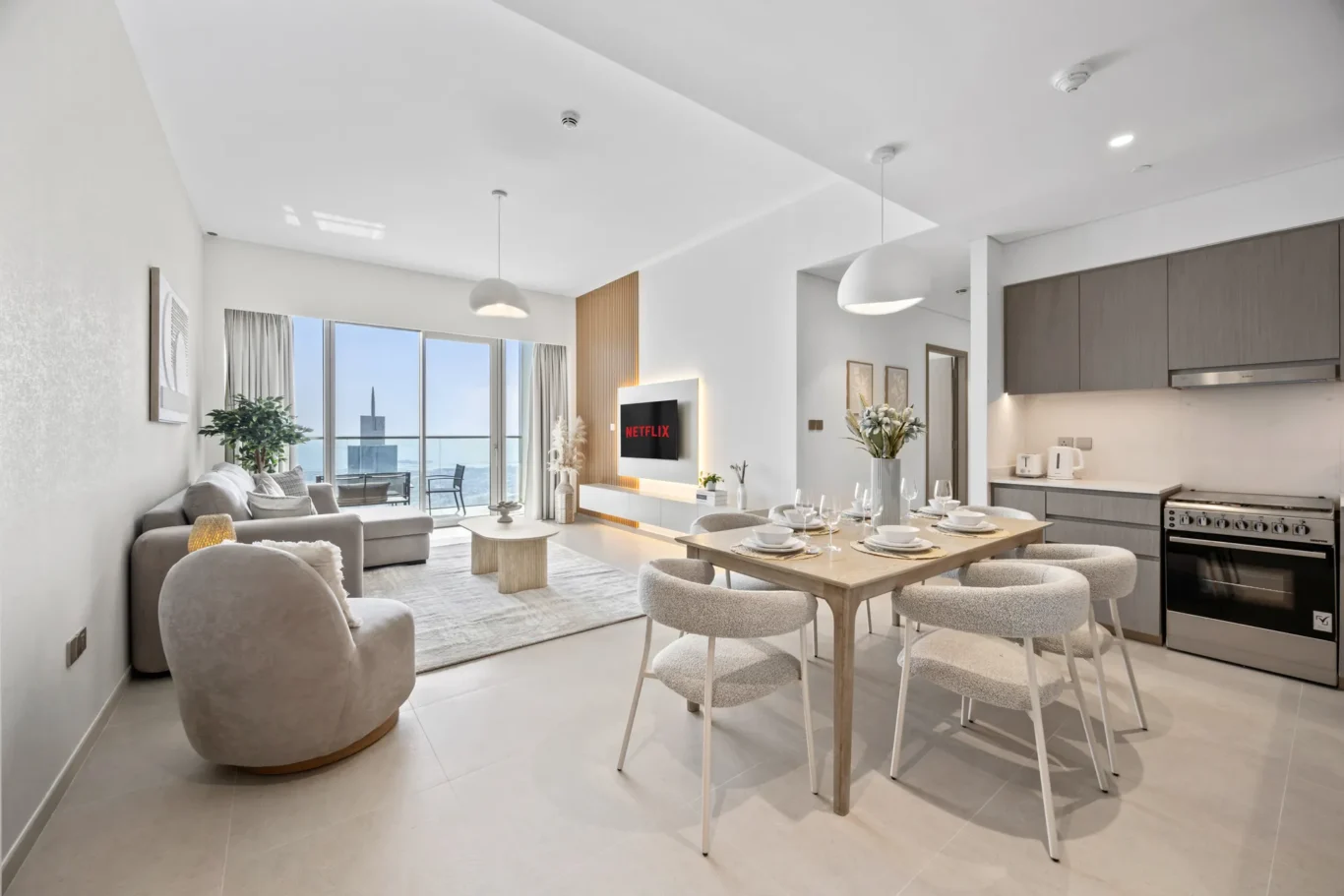 Open-plan living area with a sofa seating area, wall-mounted Netflix TV, and a dining table set for six, adjacent to a kitchen; large windows with city/ocean view.