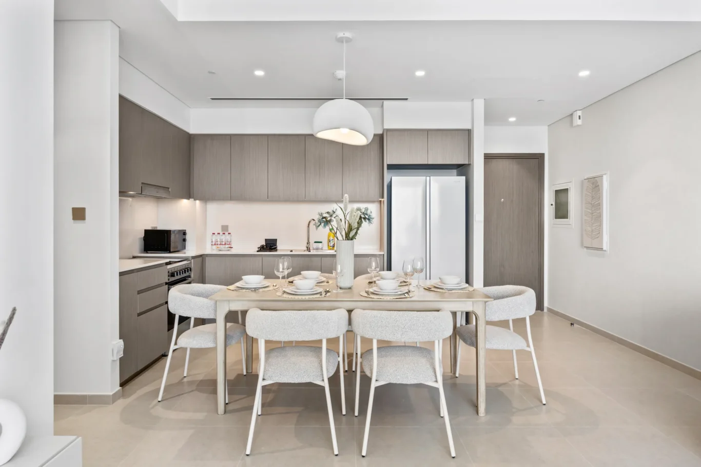 Open-plan kitchen and dining area with grey cabinets, stainless fridge, and a dining table set for six with bowls and wine glasses under a pendant light, in a bright modern space.