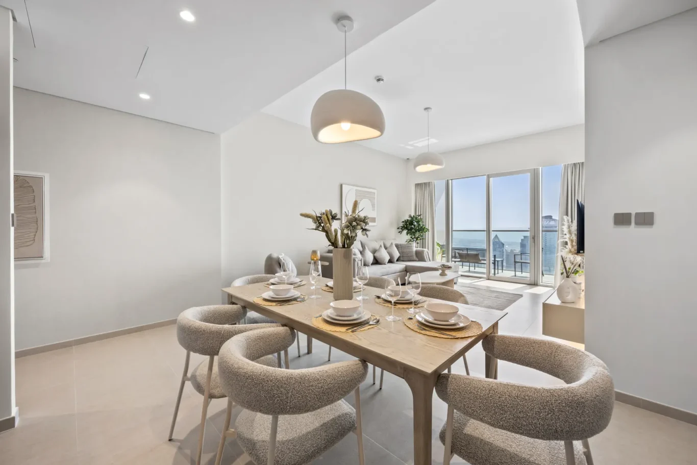 Open-concept dining and living space with a wooden table set for six, soft gray chairs, and a city-view balcony through large glass doors.