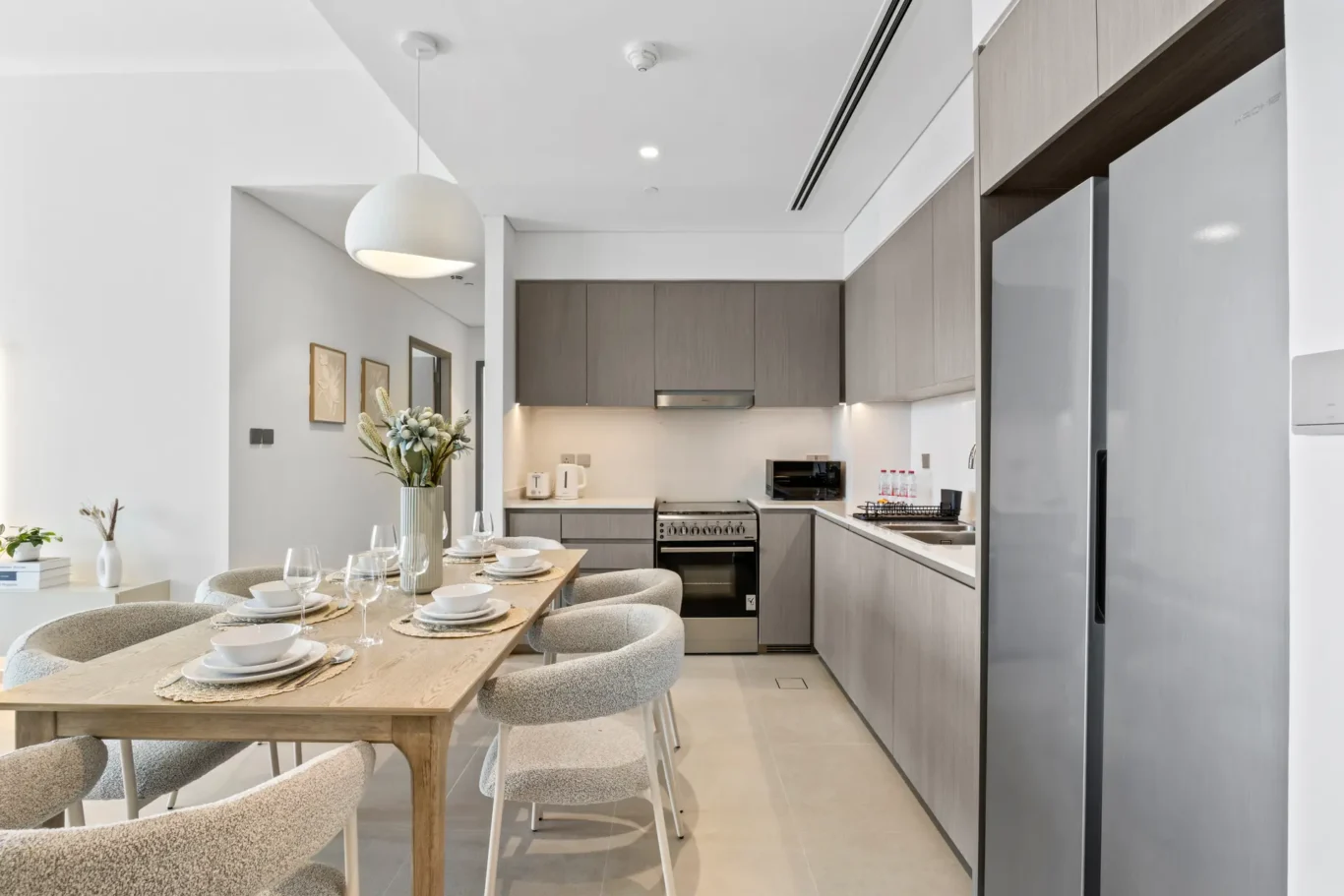 Modern open-plan kitchen with grey wood cabinets, stainless appliances, and a light wood dining table set for six with place settings and glasses.