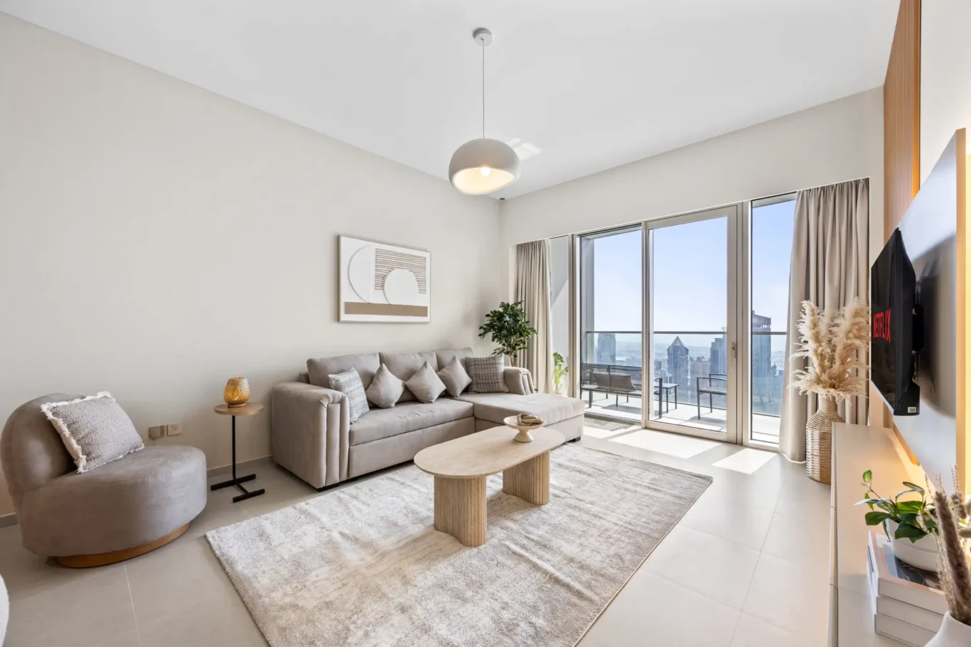 Bright living room with a beige sectional sofa, cushions, and a round coffee table by floor-to-ceiling windows showing a city skyline on the balcony.