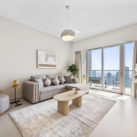 Bright living room with a beige sectional sofa, cushions, and a round coffee table by floor-to-ceiling windows showing a city skyline on the balcony.
