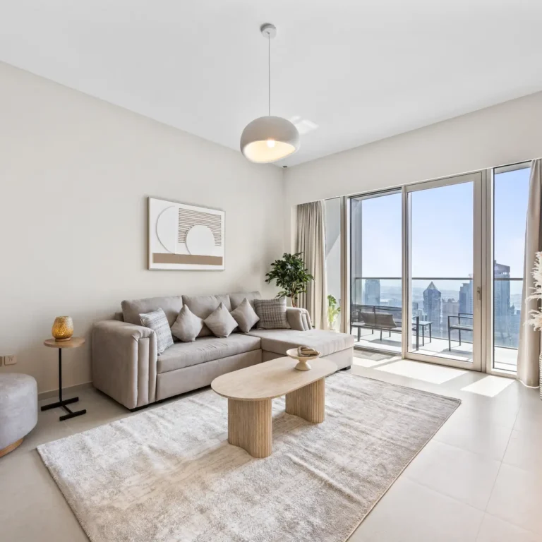 Bright living room with a beige sectional sofa, cushions, and a round coffee table by floor-to-ceiling windows showing a city skyline on the balcony.