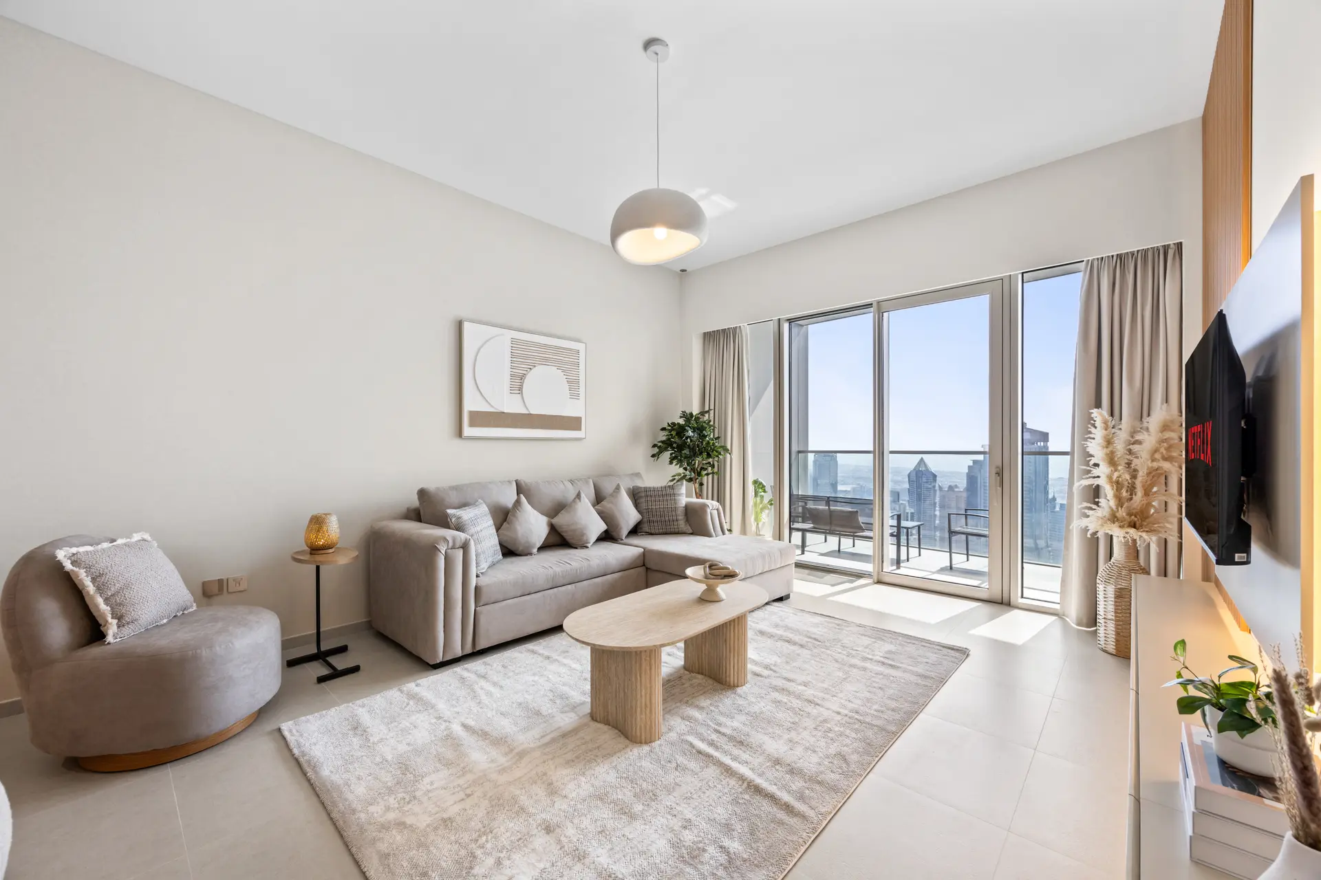 Bright living room with a beige sectional sofa, cushions, and a round coffee table by floor-to-ceiling windows showing a city skyline on the balcony.