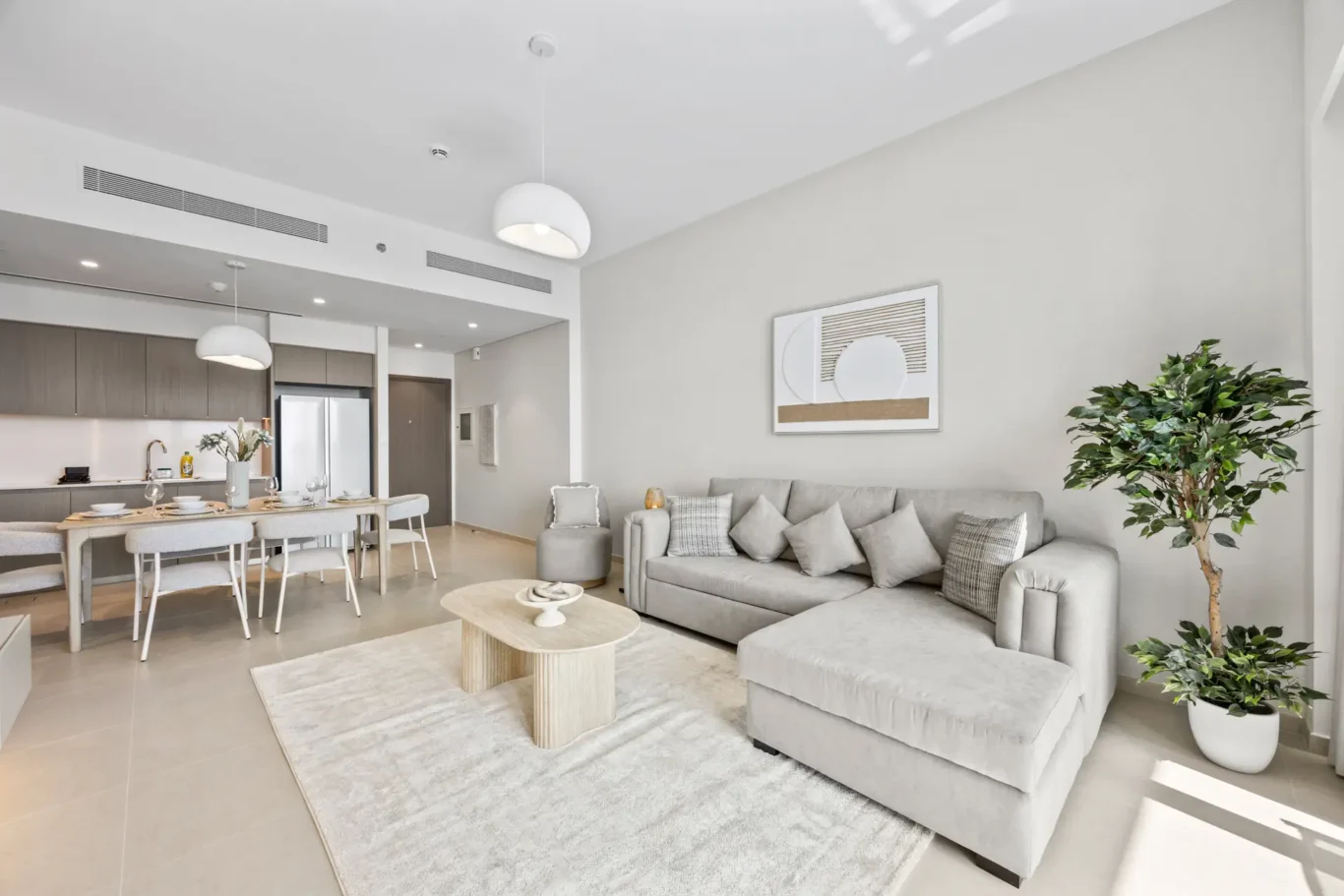 Open-concept living area with a gray sectional sofa, light wood coffee tables, a soft rug, and a tall plant in a white pot on the right side.