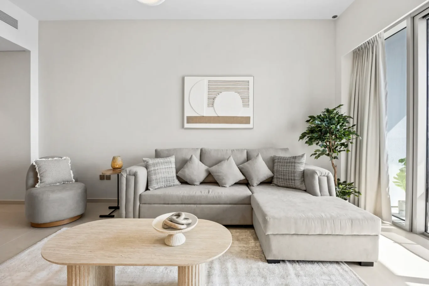 Modern living room with a light gray sectional sofa, matching cushions, and a round wooden coffee table by a large window with beige curtains and a tall plant nearby.
