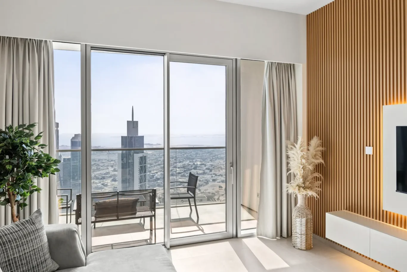 Living room with glass sliding doors opening to a balcony and a city skyline view, plus a grey sofa and wooden slat wall.