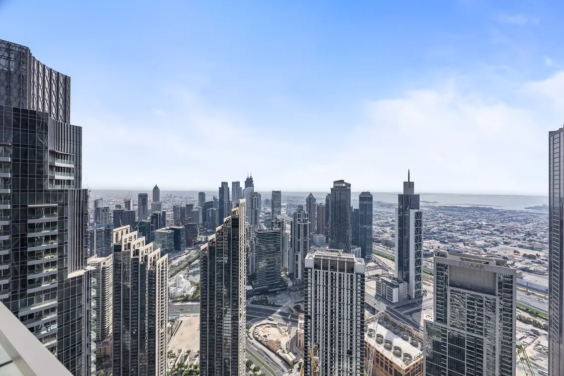 Aerial view of a dense cluster of modern glass skyscrapers forming a coastal city skyline with a distant water horizon under a clear blue sky.