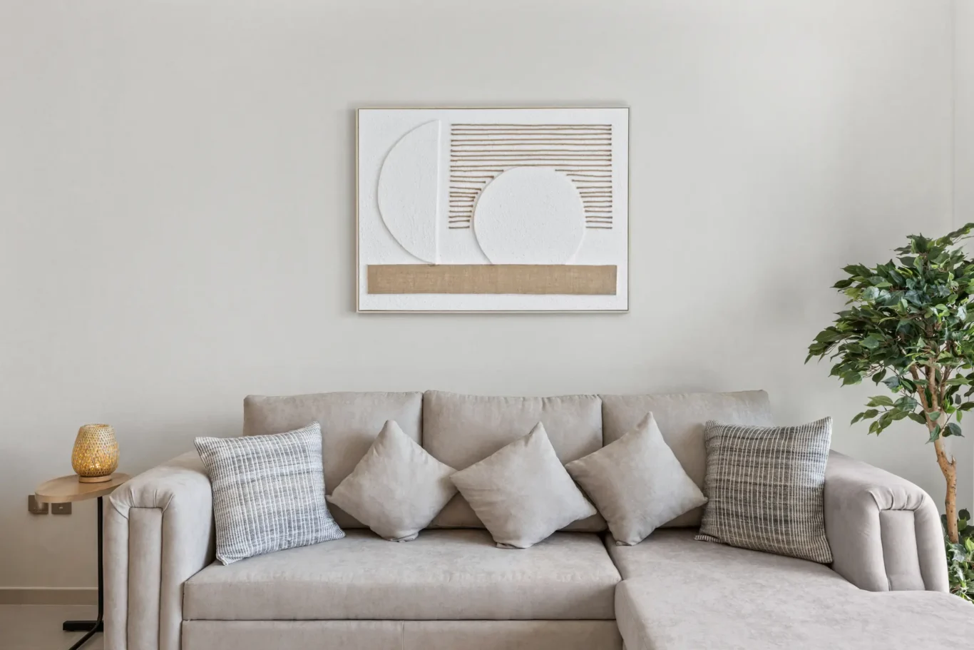 Beige sectional sofa with assorted pillows, abstract white wall art, and a potted plant on the right in a minimalist living room setting.