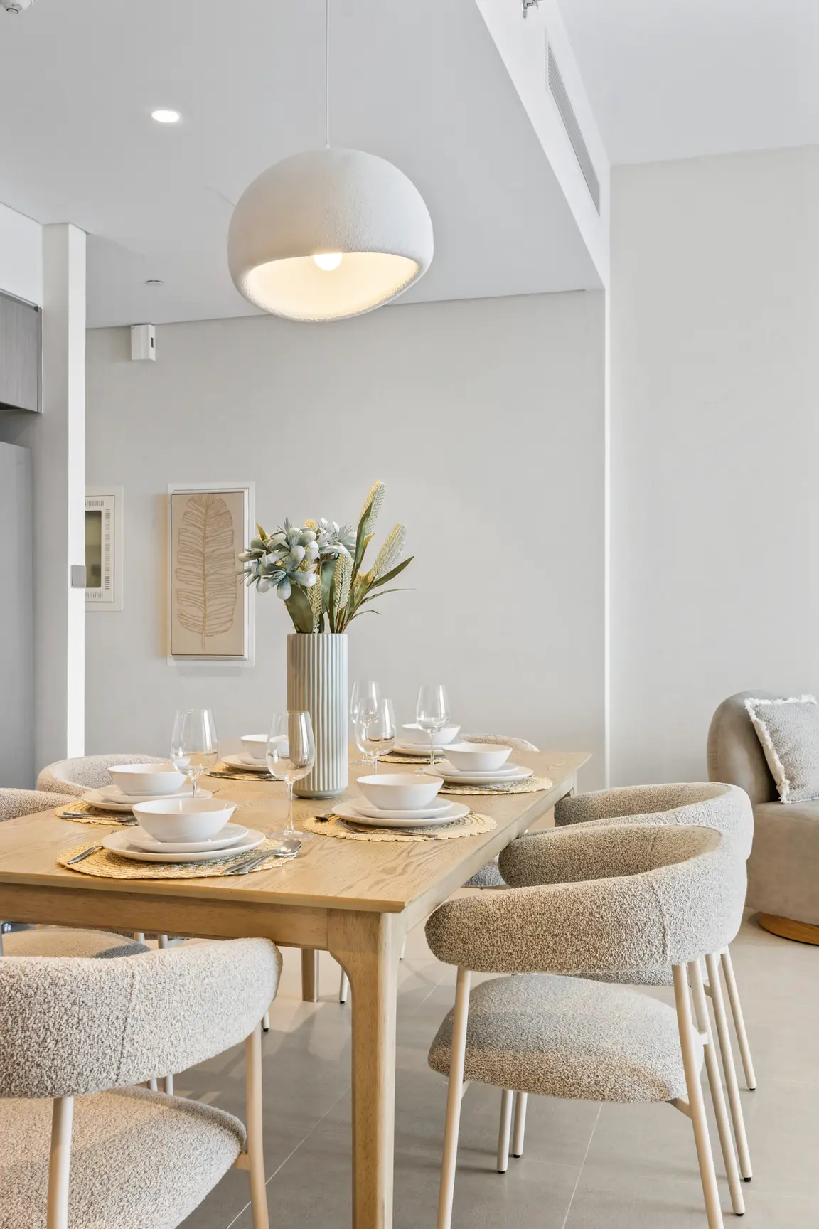 Dining room with a light wood table set for six, beige boucle chairs, and a tall ribbed vase of greenery.
