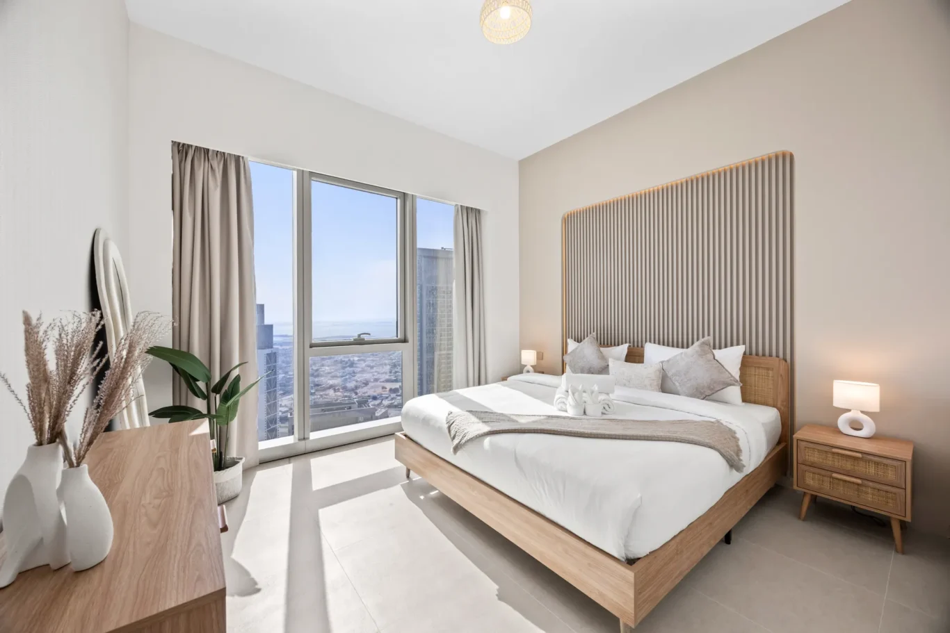 Bright hotel bedroom featuring a wooden platform bed with a tall, striped headboard, two nightstands, and large windows with beige curtains offering a city view by day.