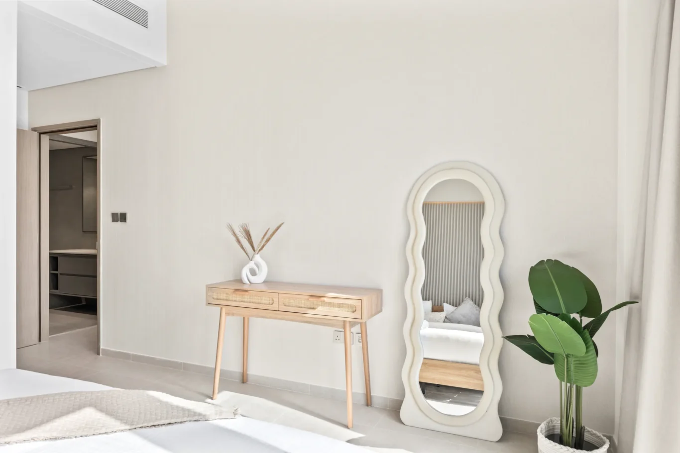Minimalist bedroom corner with a wavy-edged full-length mirror, a light wood console table, and a potted plant by beige walls.