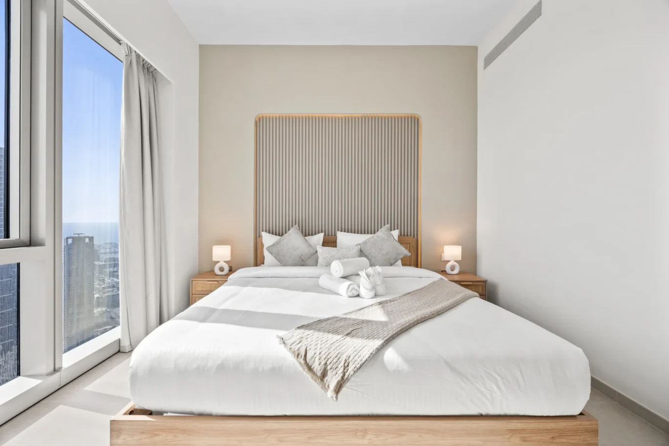 Modern hotel-style bedroom: platform bed with white linens, tufted beige headboard with warm backlighting, and matching nightstands with lamps near large windows showing a city skyline.