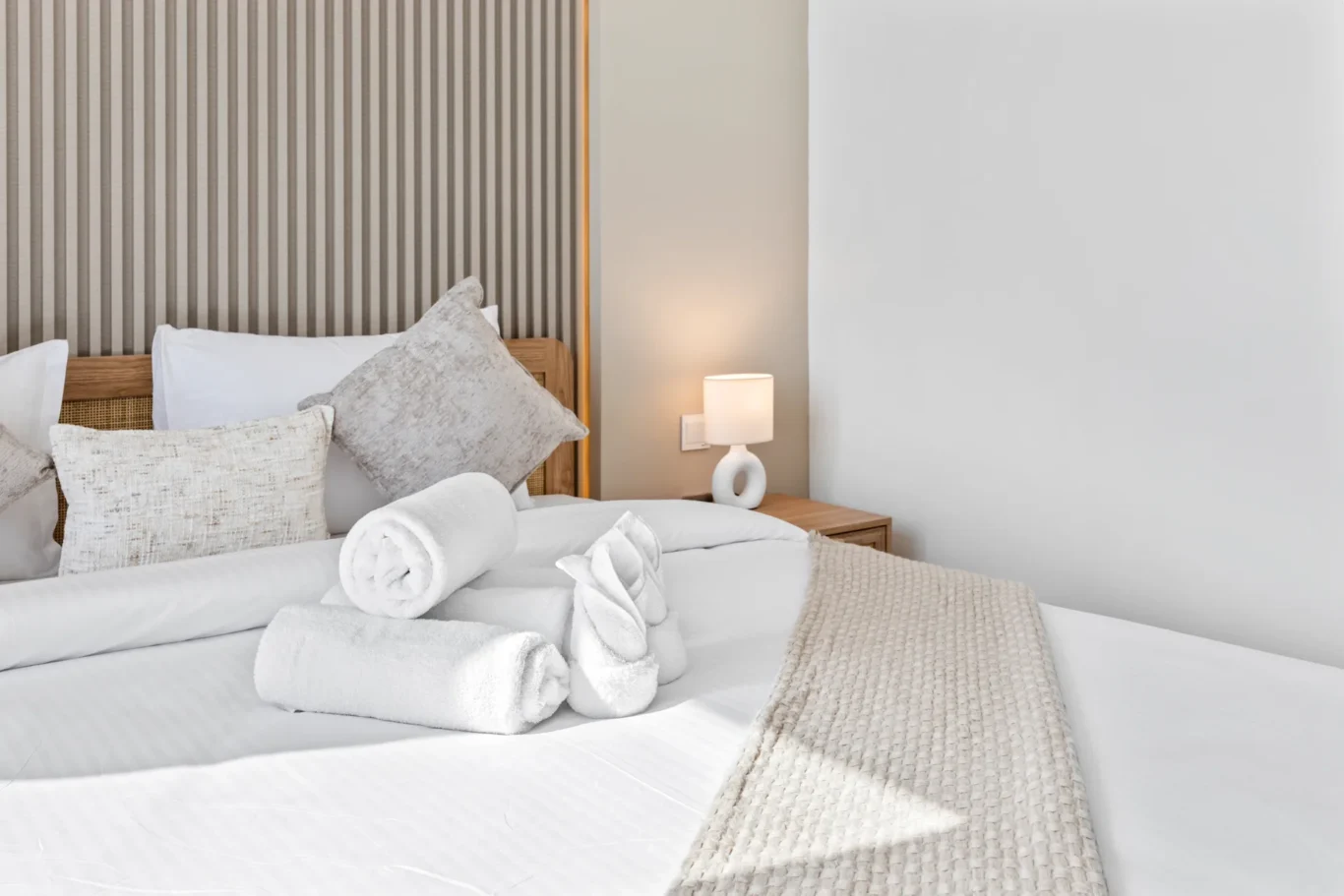 Neatly made bed with white bedding, beige knitted throw, decorative pillows, and two rolled white towels; bedside lamp glows on a wooden nightstand by a striped headboard panel.