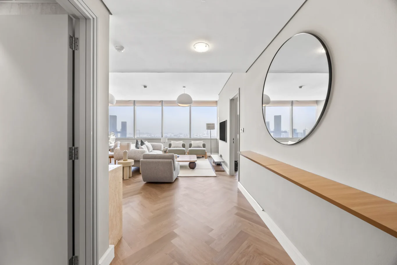 Wide view of a bright modern living room with floor-to-ceiling city-view windows and a large circular mirror on the right wall reflecting the space.