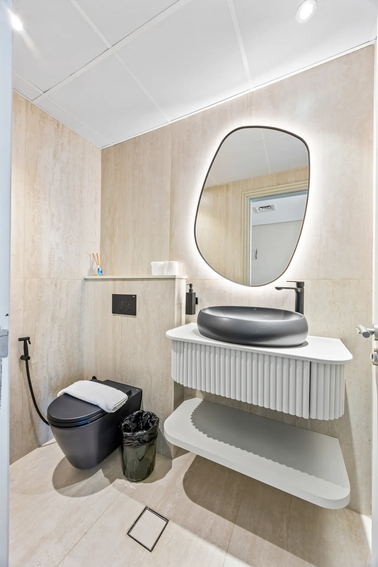 Beige-tiled modern bathroom with a black vessel sink, backlit oval mirror, and wall-mounted vanity.