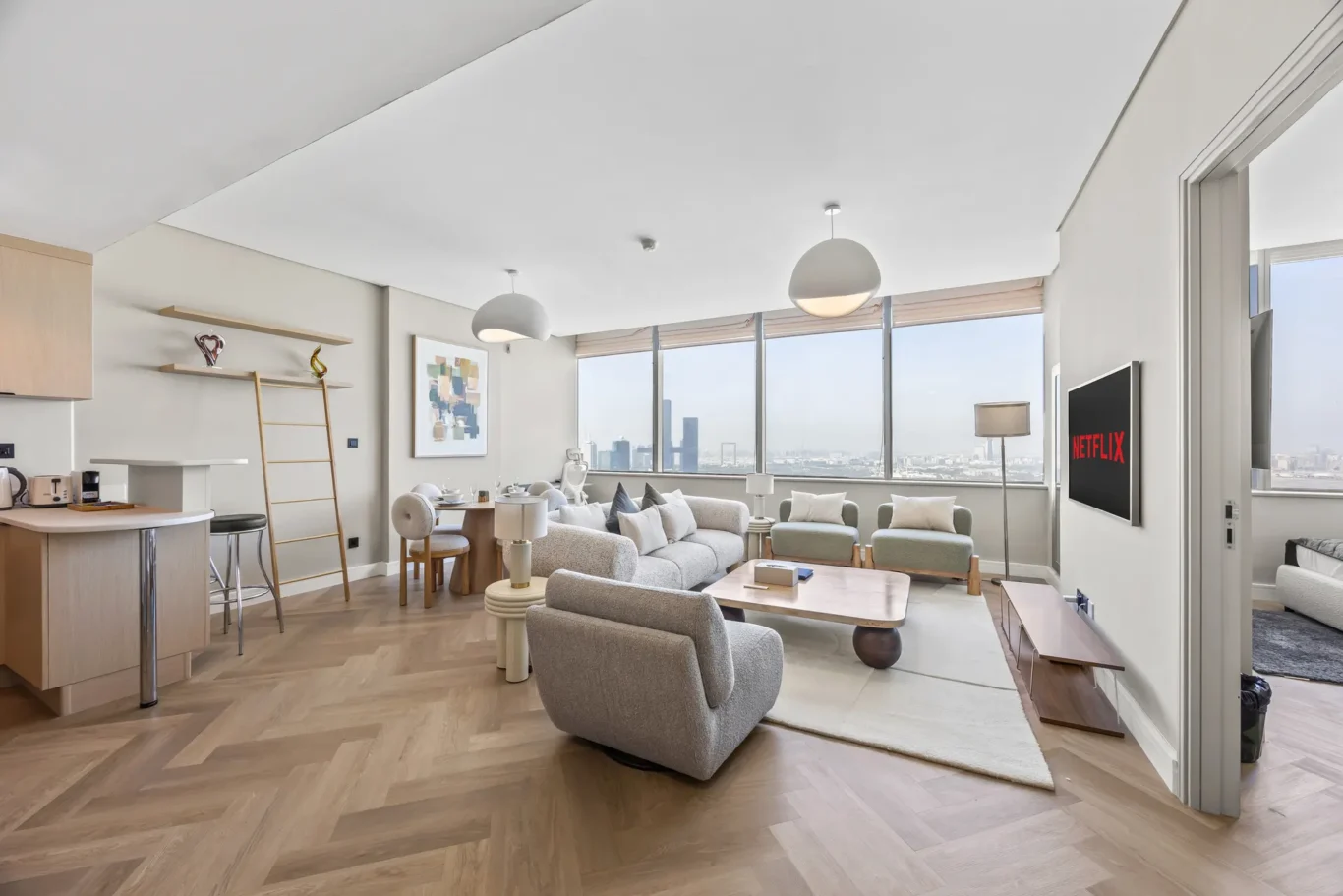 Bright open-plan living area with a city skyline view through large windows, cream sofas, and a wooden coffee table arranged around a white rug.