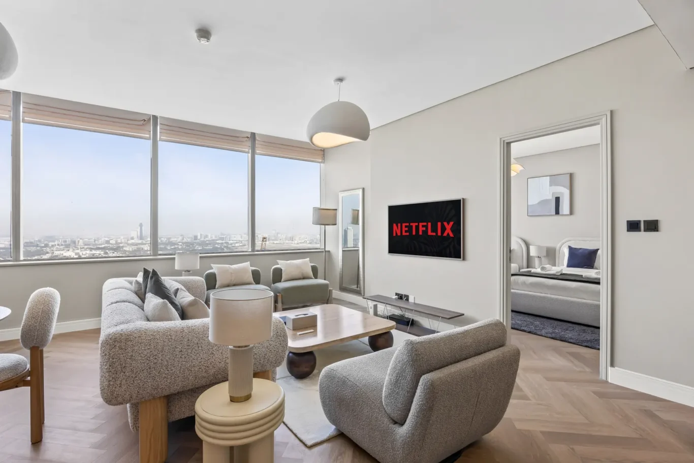 Bright modern living room with large windows, gray furniture, and a Netflix screen on the wall beyond a coffee table.”/“Bright modern living room with large windows, gray furniture, and a Netflix screen on the wall.