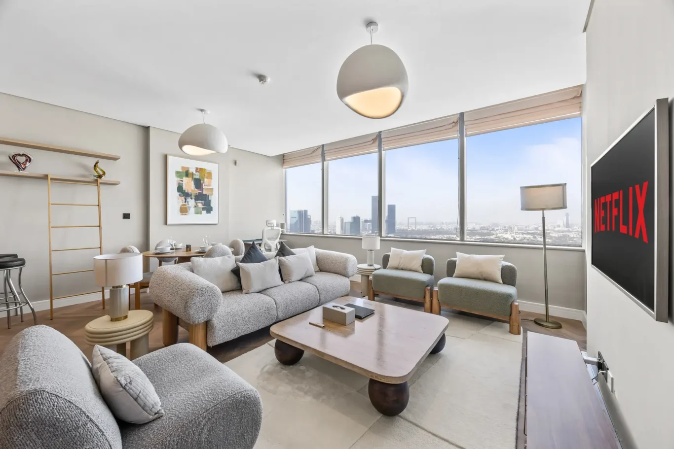 Modern living room with large windows and a city skyline view, gray sofa set, and a Netflix screen on the wall to the right.
