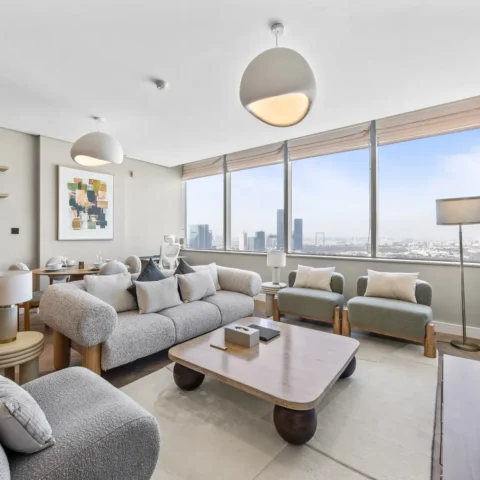Modern living room with large windows and a city skyline view, gray sofa set, and a Netflix screen on the wall to the right.