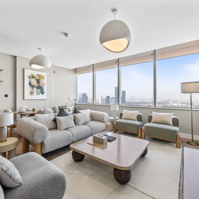Modern living room with large windows and a city skyline view, gray sofa set, and a Netflix screen on the wall to the right.
