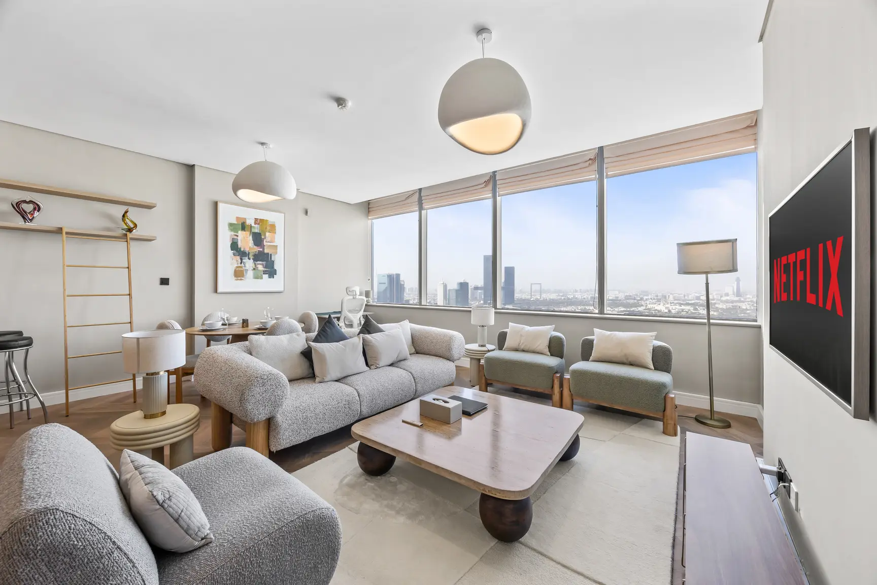 Modern living room with large windows and a city skyline view, gray sofa set, and a Netflix screen on the wall to the right.