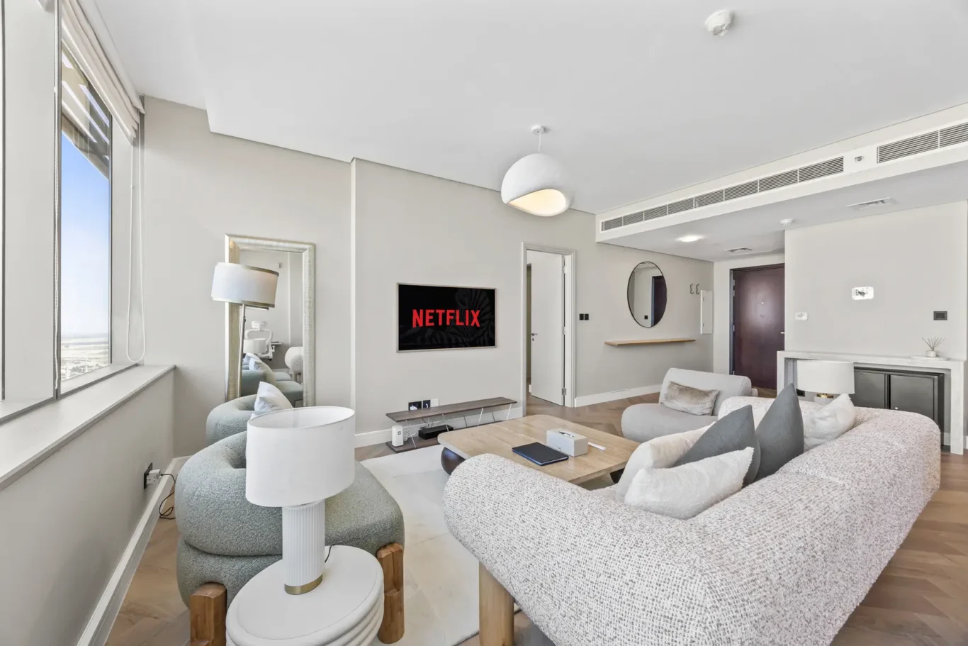 Bright modern living room with a wall-mounted Netflix screen and gray sofas facing it. large windows on left let in natural light.