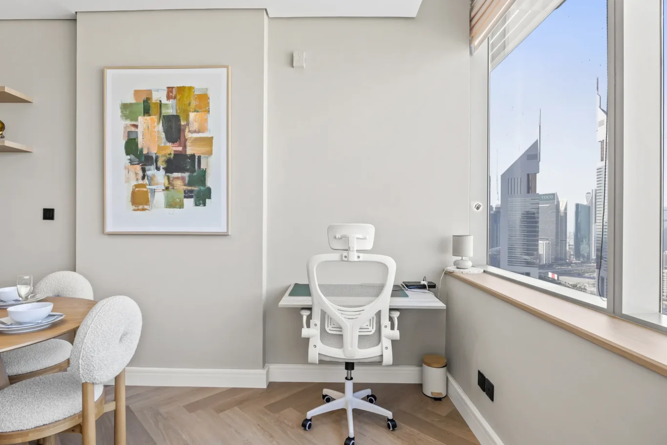 Bright home office with a white ergonomic chair at a small desk by a large window overlooking a city skyline; abstract painting on the wall to the left.